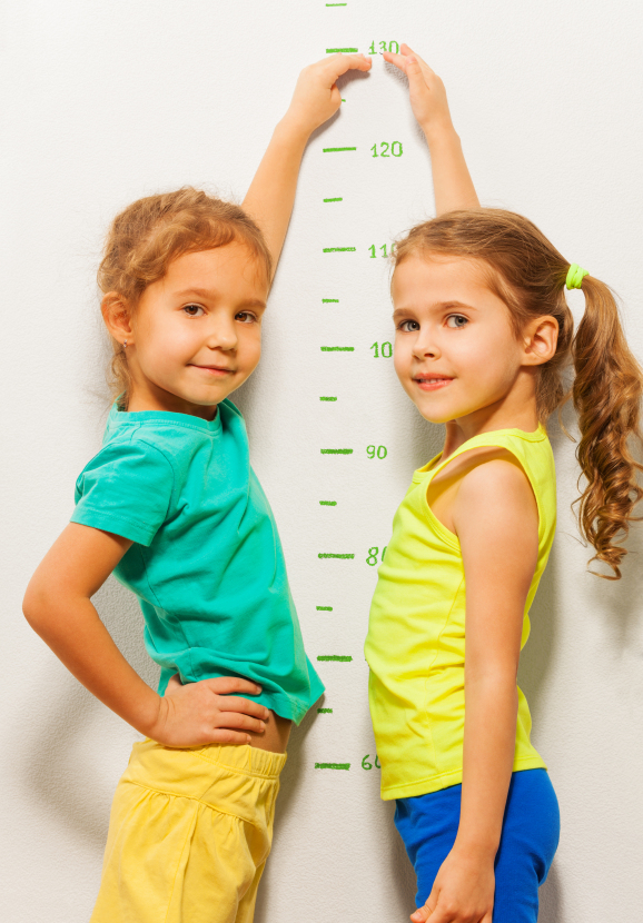 Two girls show height on the scale pretending to grow faster looking at camera and smiling