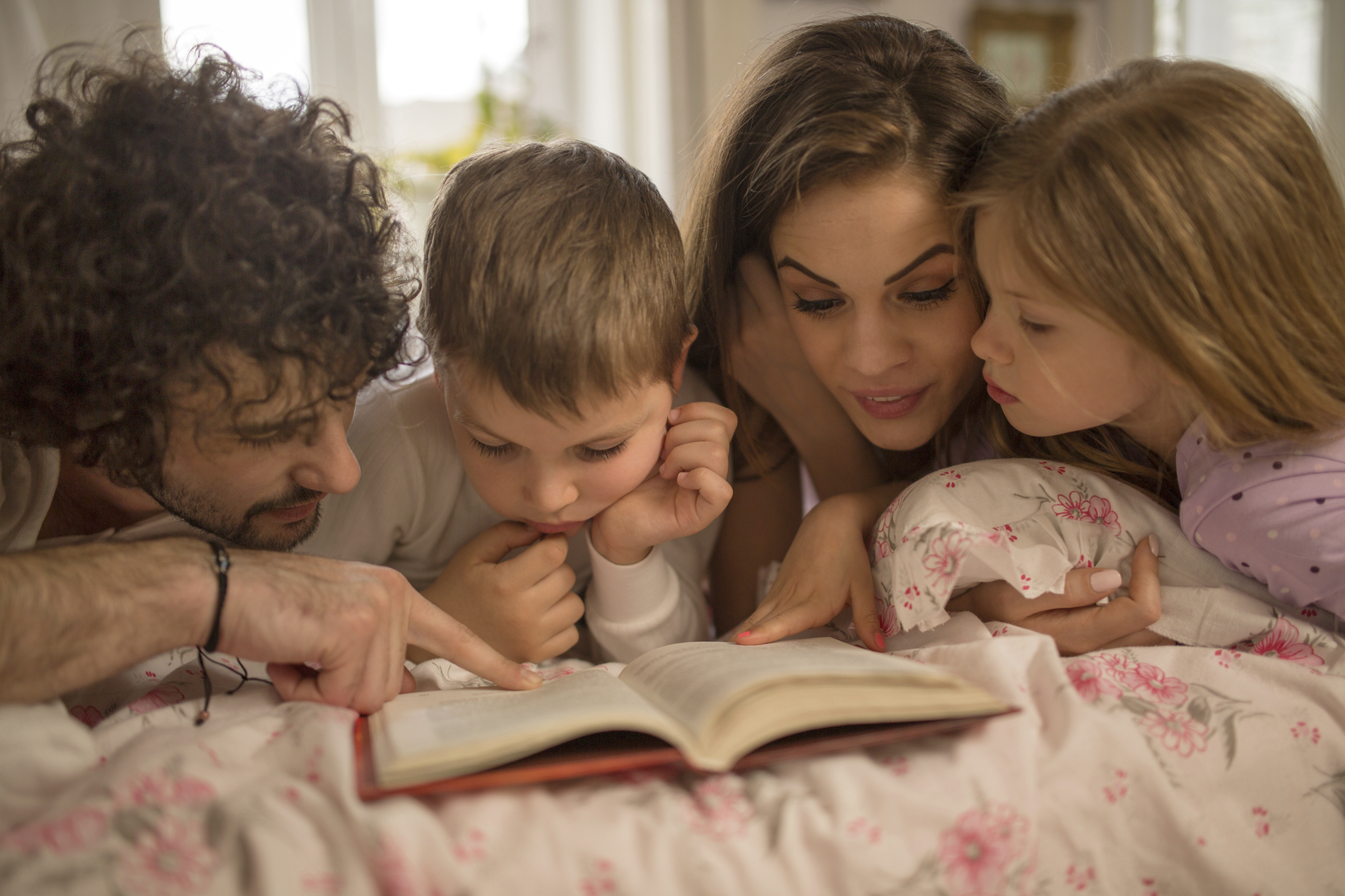 Young parents reading book together with their children in bedroom.