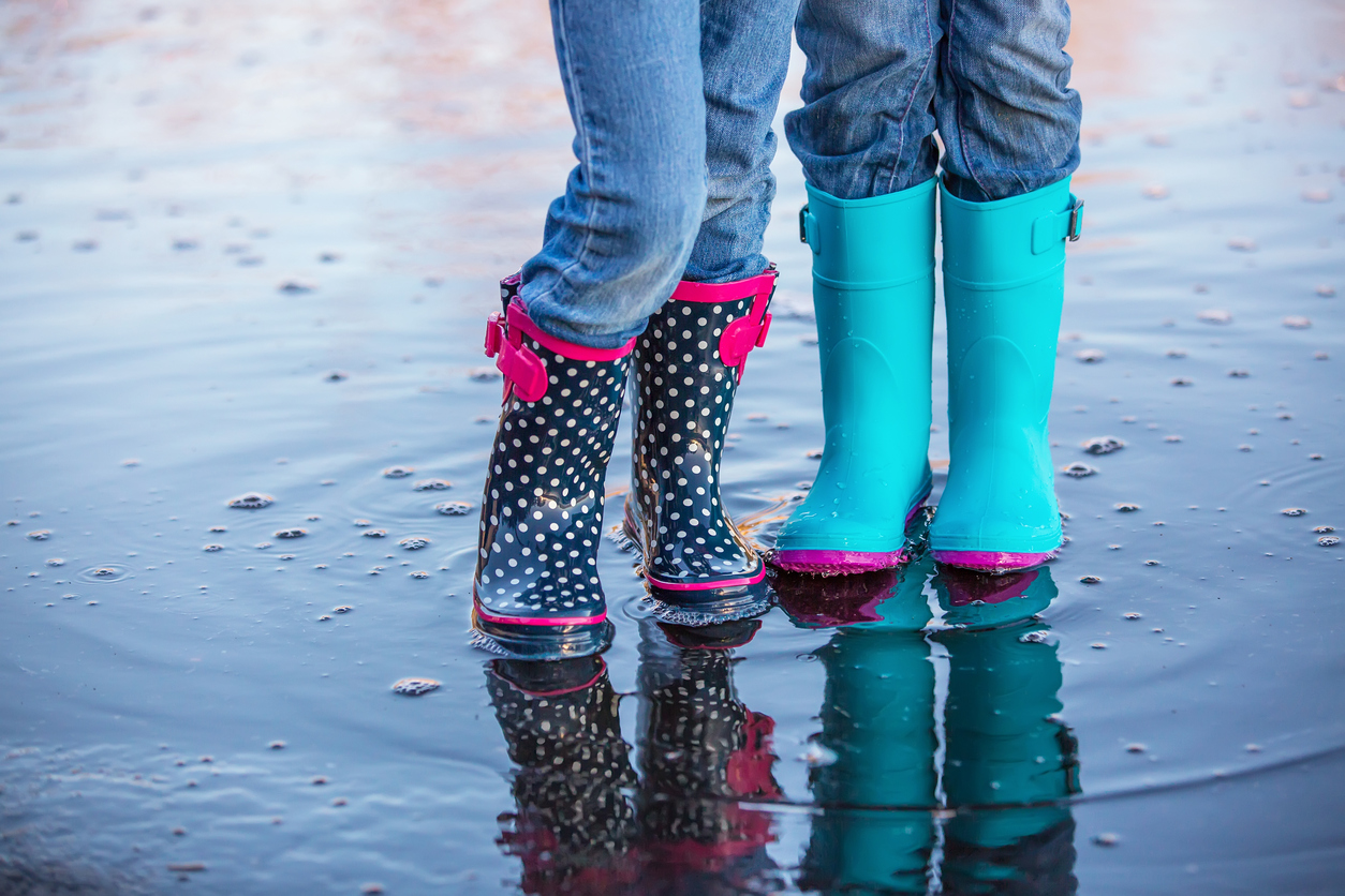 Waist down view of two children wearing rubber boots while standing side by side in a springtime water puddle. Both girls are wearing light blue denim jeans. The girl on the left has navy blue with white polka dots and pink trim on her boots. The girl on the right has light blue boots with pink bottoms.